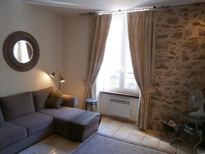 Ein Wohnzimmer mit einer Couch, einem kleinen Tisch, einem Standventilator, einem Heizgerät und einem Fenster mit Vorhängen.