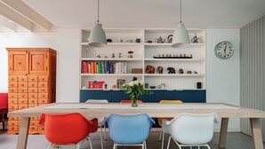 Ein Essbereich mit einem Holztisch, bunten Stühlen, einem Bücherregal und einer Uhr an der Wand.