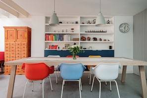 Ein Essbereich mit einem Holztisch, bunten Stühlen, einem Bücherregal und einer Uhr an der Wand.