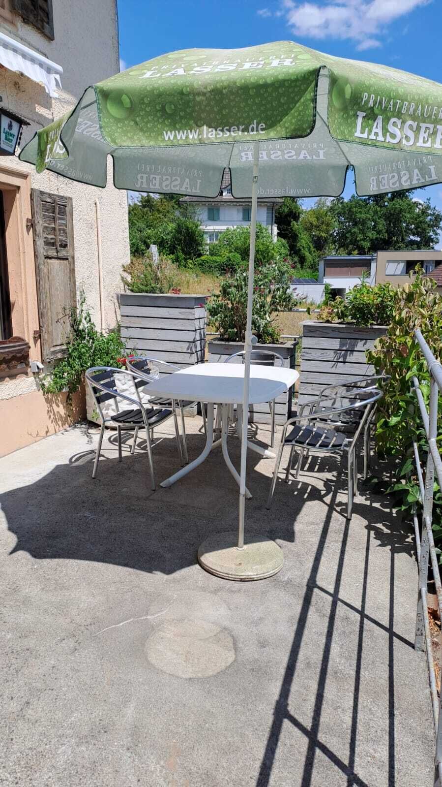 An outdoor patio with a table and chairs under a green umbrella, surrounded by greenery and buildings.