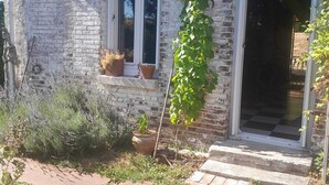 En rustik stenbyggnad med ett stort fönster, en dörr och en liten veranda med krukväxter.