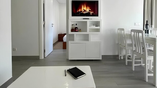 A modern living room with a flat-screen TV mounted above a white entertainment unit, a dining area with white chairs, and a small table with a notebook and pen. - Rocha's Corner (Portimão)