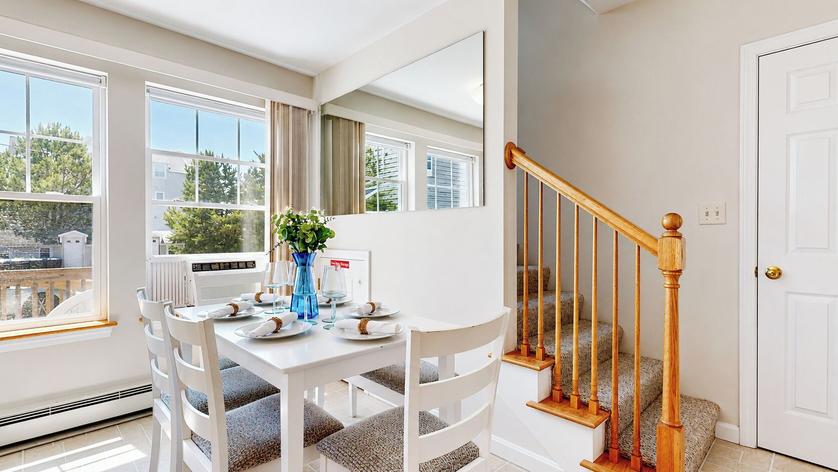 A dining area with a white table set for four, a blue vase with flowers, and a staircase with wooden railings.