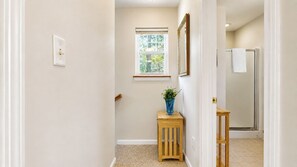 A hallway with a window, a wooden stool with a vase, and a shower area.
