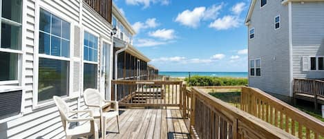Une terrasse en bois offrant une vue sur la plage, un bâtiment blanc doté de grandes fenêtres et un ciel bleu azur.