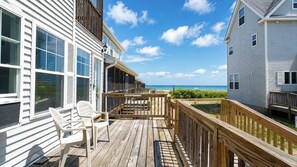 A wooden deck with a view of the beach, a white building with large windows, and a clear blue sky.