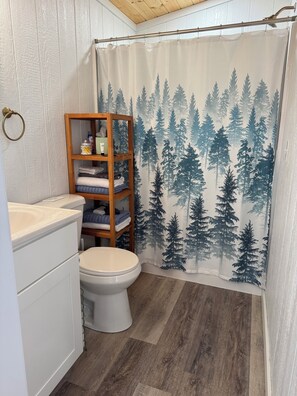 A bathroom with a shower curtain featuring a forest pattern, a wooden shelf with towels, and a white toilet. - Liberty-Cabin-2 @ Eagle Hollow (goreville)