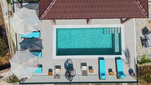 An aerial view of a rectangular swimming pool with a tiled deck surrounding it, equipped with multiple lounge chairs and umbrellas.