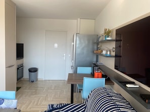 A compact kitchen with a refrigerator, stove, and a trash bin. There is a dining area with a table and chairs, and a sofa with a striped pillow. - Cannes Marina Studio with Harbor View, Pool (Mandelieu-la-Napoule)