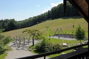 Ein Spielplatz mit Schaukeln und Klettergerüsten, umgeben von einer Rasenfläche und Bäumen, Blick von einem überdachten Balkon.