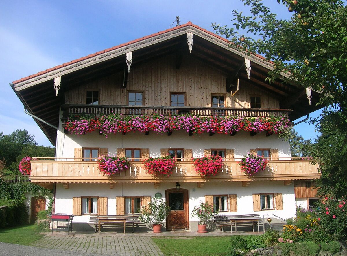 A two-story wooden building with a balcony adorned with flowers, a front entrance, and a paved area with benches.
