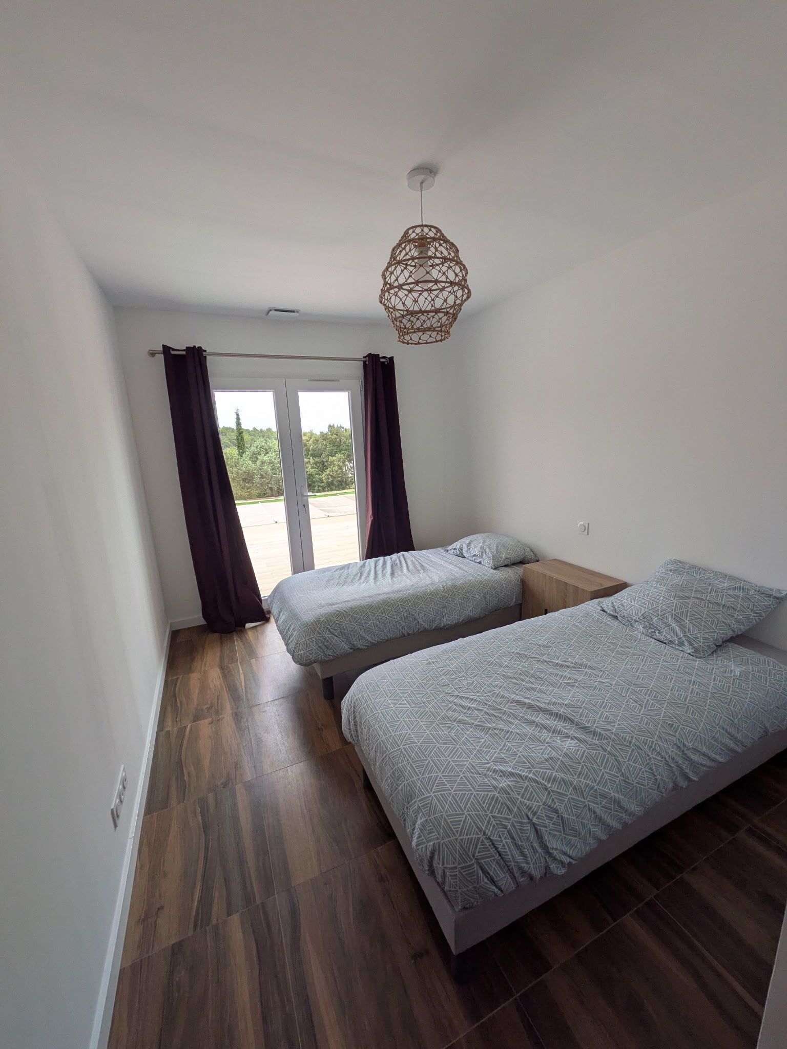A room with two beds, a ceiling light fixture, and a view through a door to an outdoor area.