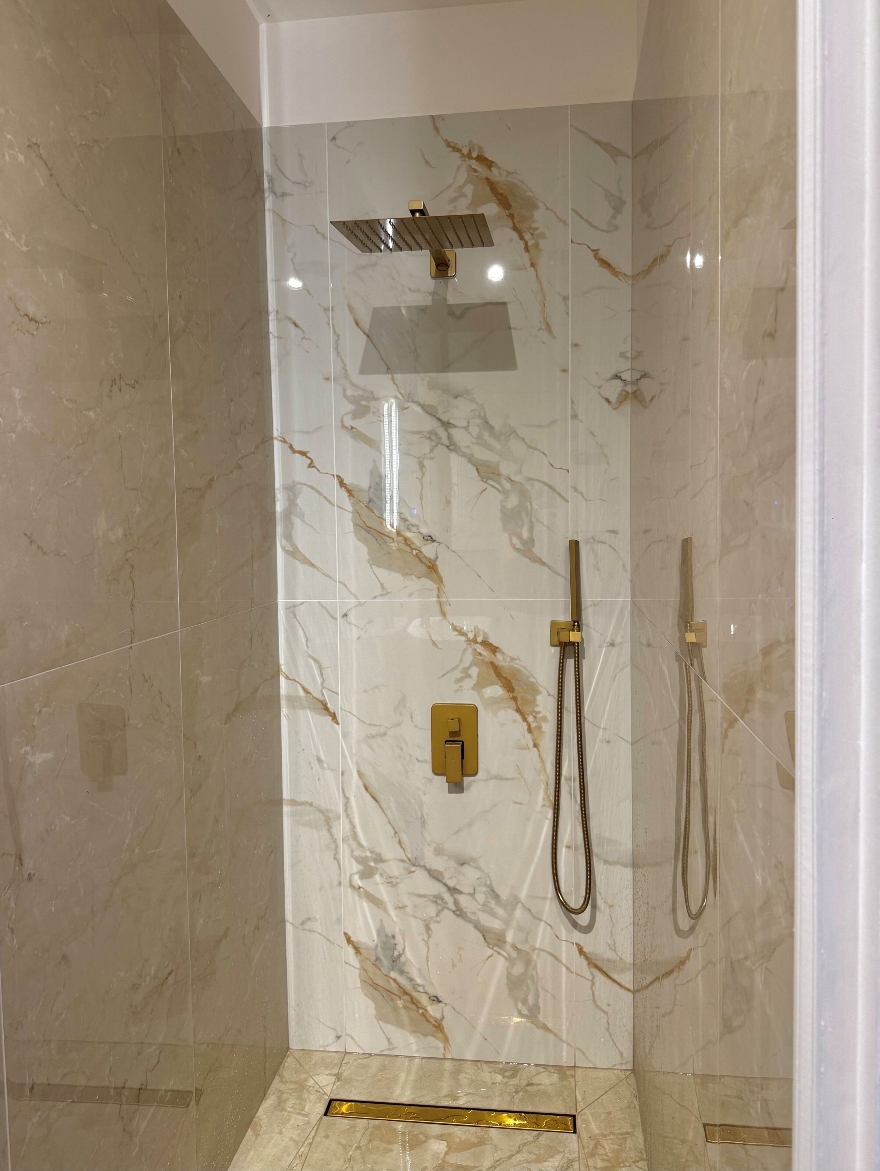 A glass-enclosed shower with a marble wall, a handheld showerhead, and a wall-mounted control panel.