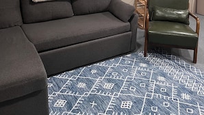 Ein Zimmer mit Ecksofa, einem grünen Ledersessel, einem gemusterten Teppich und einem Fenster mit Blick auf Bäume.