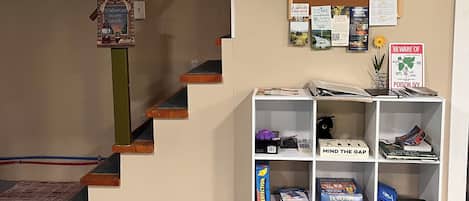 A staircase with a wooden handrail, a bulletin board with various notices, and a white shelving unit with books and games.