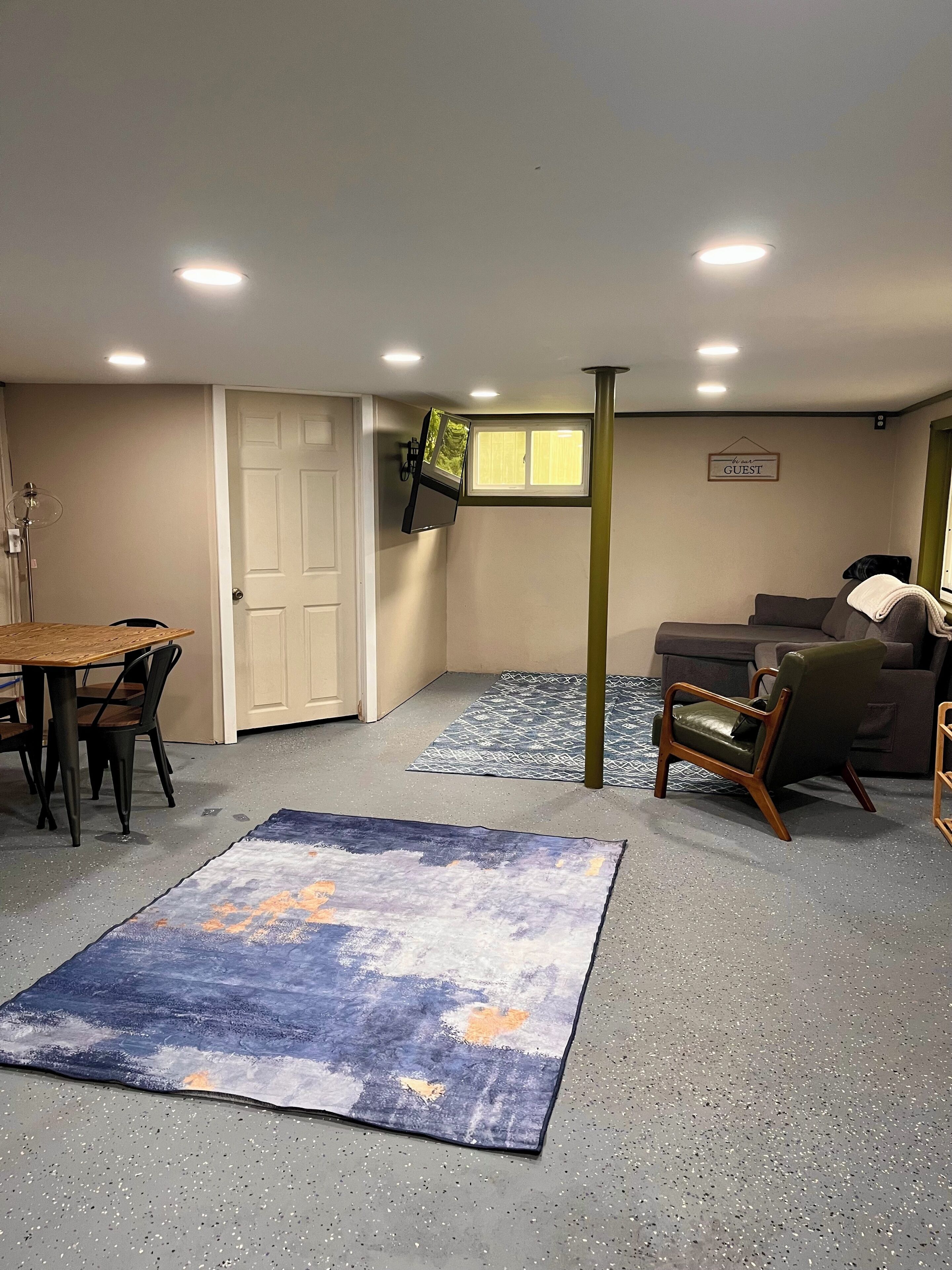 A room with a dining table, chairs, a sofa, a TV mounted on the wall, and a rug on the floor.