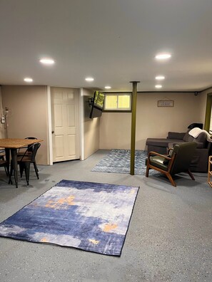 A room with a dining table, chairs, a sofa, a TV mounted on the wall, and a rug on the floor.