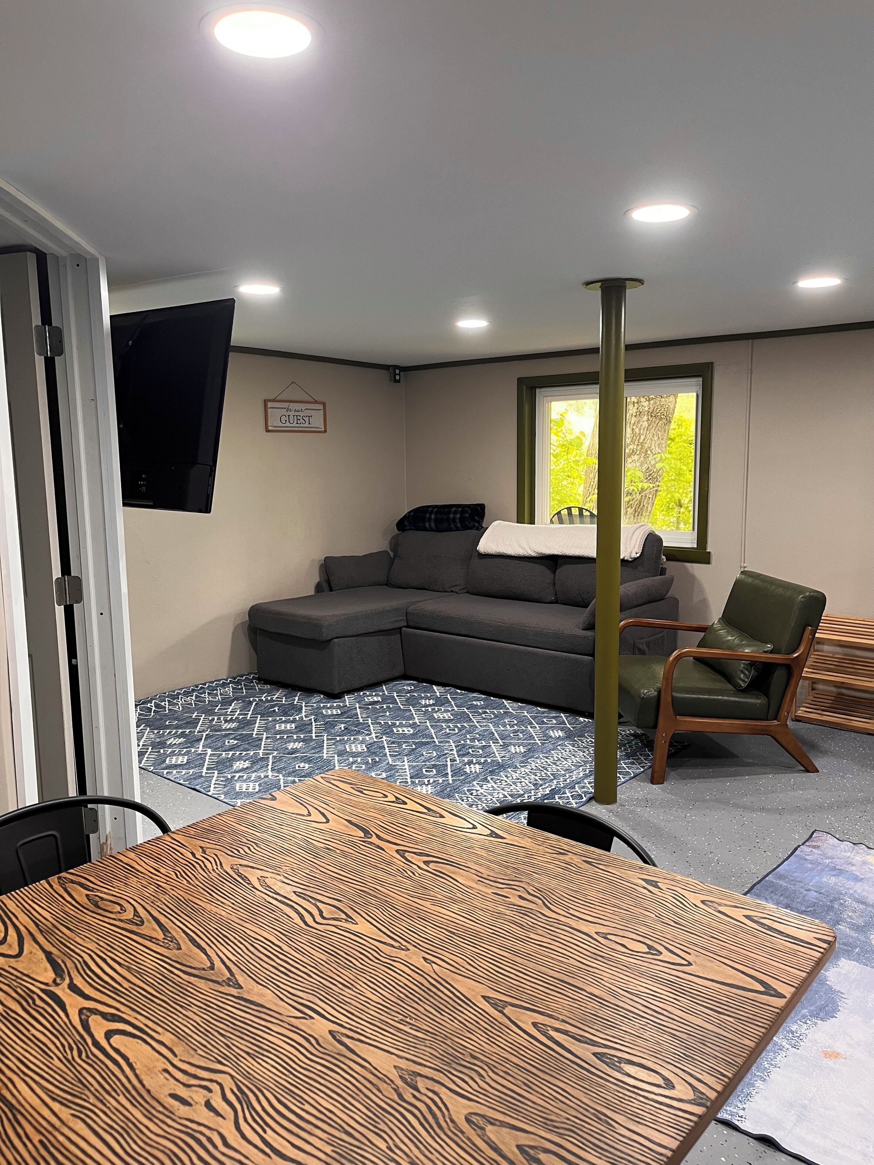 Une pièce comprenant un canapé d’angle, une table à manger en bois, un fauteuil vert et une fenêtre donnant sur un paysage verdoyant.