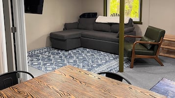 Une pièce comprenant un canapé d’angle, une table à manger en bois, un fauteuil vert et une fenêtre donnant sur un paysage verdoyant.