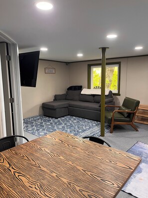 Una habitación con sofá modular, una mesa de comedor de madera, un sillón verde y una ventana con vistas a zonas verdes.