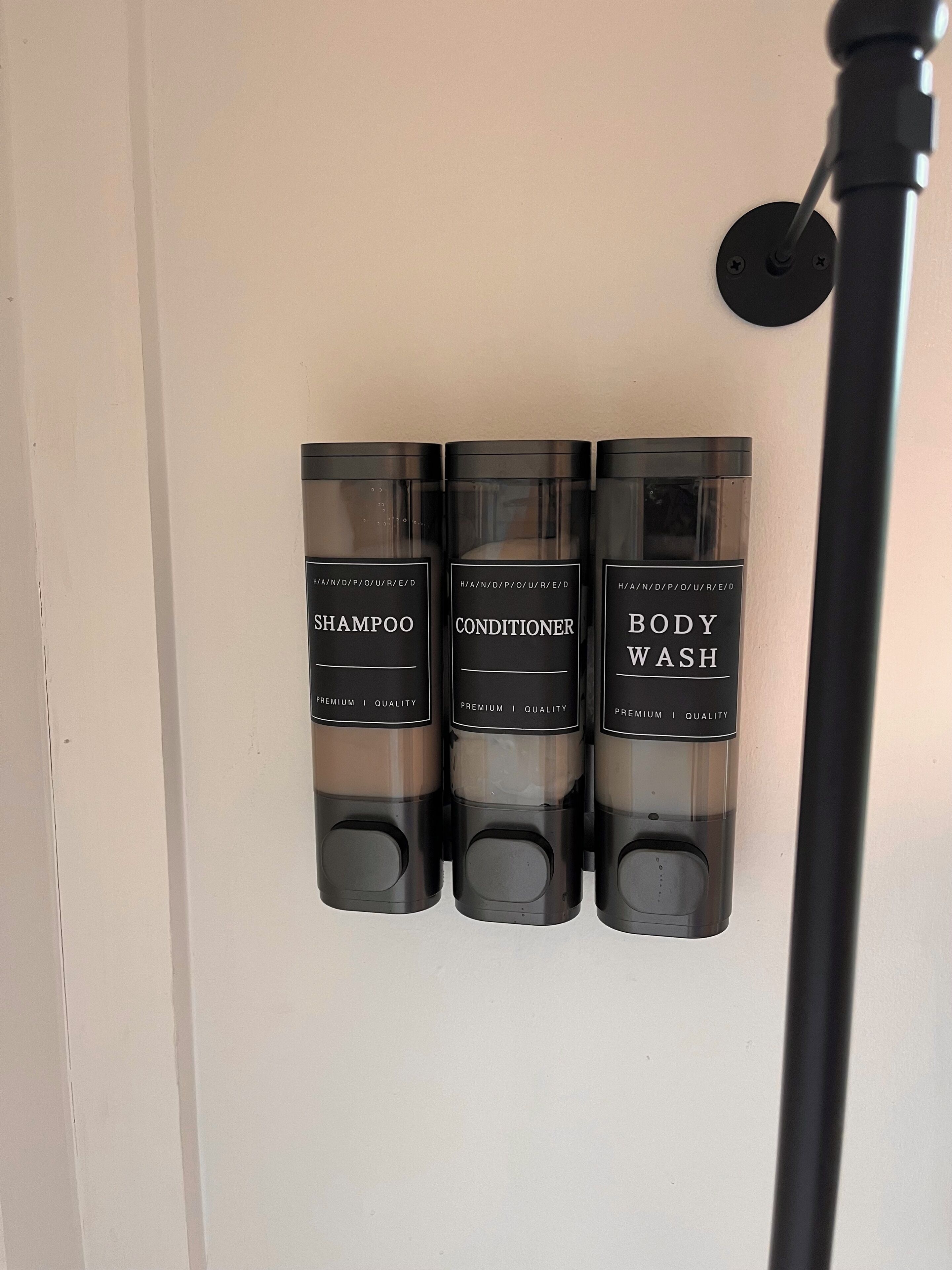 Tres botellas negras etiquetadas 'SHAMPOO', 'CONDITIONER' y 'BODY WASH' fijadas en una pared.