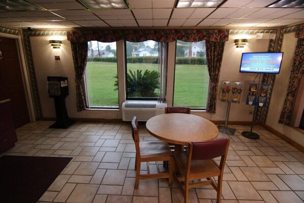 A hotel room with a dining table, chairs, a television, and a view of a lawn through large windows.