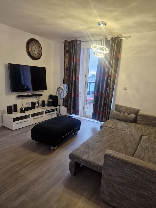 A living room with a flat-screen TV, a white entertainment unit, a black ottoman, a grey sofa, and a ceiling fan. - Wembley View Apartment (London)