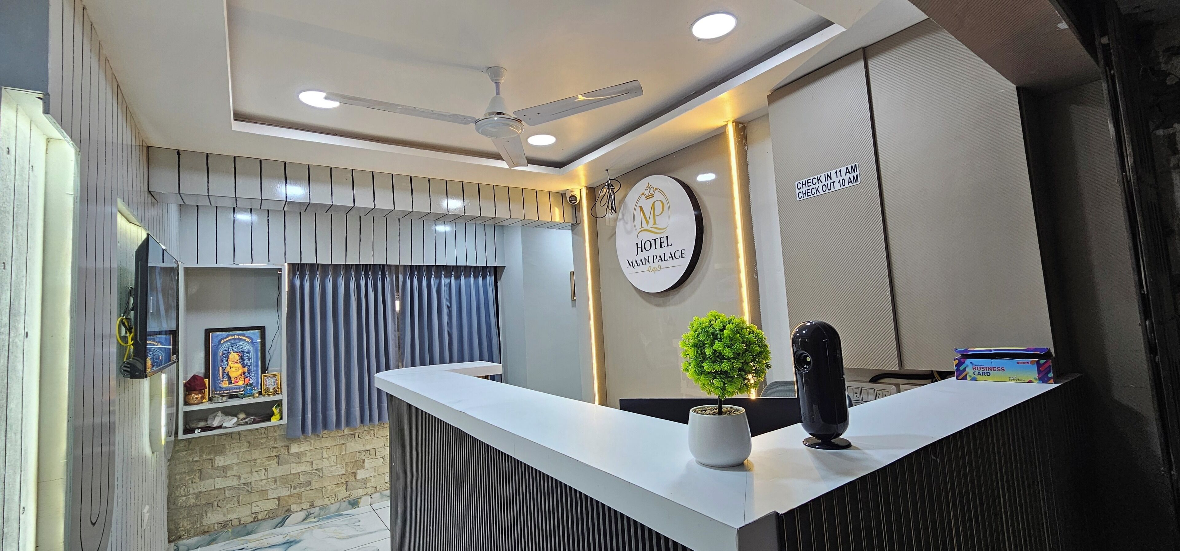 A hotel reception area with a modern design, including a reception desk, a ceiling fan, a wall-mounted sign, and a small potted plant.
