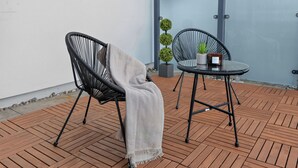 Eine Terrasse mit zwei schwarzen Korbstühlen, einem runden Tisch und einem kleinen Blumentopf.