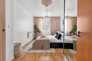 A modern bedroom with a bed, bedside tables, a radiator, a window with curtains, and a potted plant. - Modern 2 Bed 2 Bath Stay | Stratford, Olympics Park, ABBA Voyage &City / Sleep 5 (Stratford)