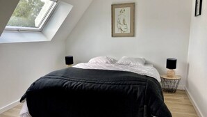 Un dormitori amb sostre inclinat, un llit amb mantell negre, almoines blanques, dues làmpades laterals negres, una obra d'art amb marc a la paret i una taula rodona petita amb una làmpada.