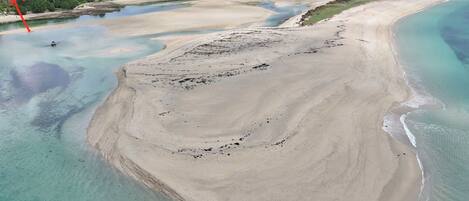 An aerial view of a coastal area with a large sandy beach, a river, and a forested area in the background.
