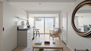 Eine moderne Küche mit Mikrowelle, Wasserkocher und Barhocker. Ein Esstisch mit Büchern und ein Korbstuhl sind ebenfalls vorhanden. An der Wand befindet sich ein großer Spiegel.