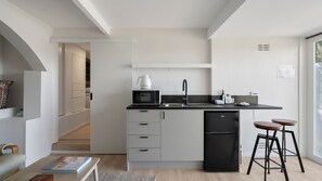 Eine moderne Küche mit einem schwarzen Kühlschrank, einer Mikrowelle und einem Wasserkocher auf der Arbeitsplatte. Zwei Hocker und eine kleine Bar laden zum Verweilen ein.