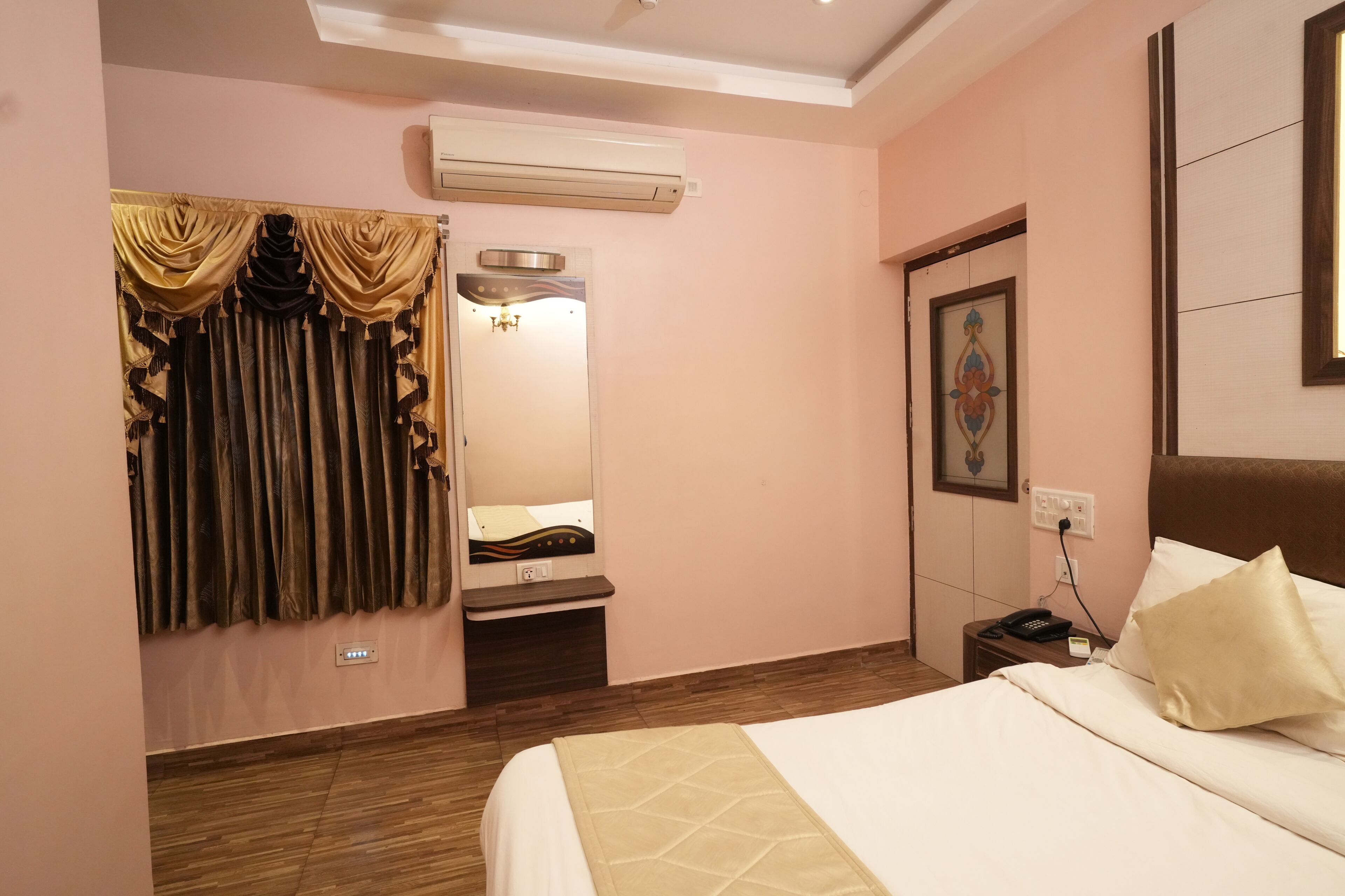 Una habitación de hotel con cama, tocador con espejo, unidad de aire acondicionado, mesita de noche y teléfono fijo en la pared.