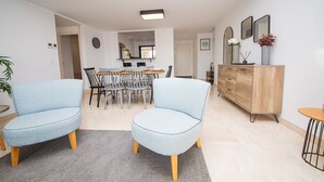 A modern living room with two light blue armchairs, a wooden dining table with chairs, a grey carpet, and a wooden cabinet with a mirror and decorative items. - Lotus Doña Julia 7.1A (Casares)