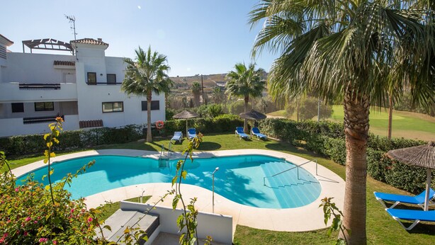 A swimming pool with a curved edge, surrounded by a grassy area with palm trees and lounge chairs. - Lotus Doña Julia 7.1A (Casares)