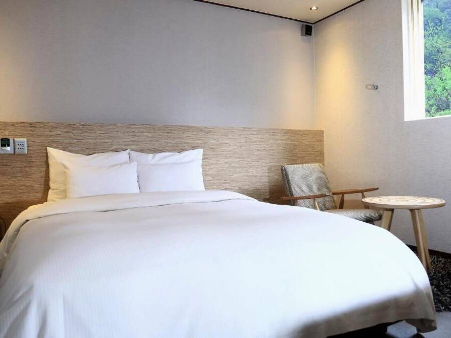 A modern hotel room with a large bed, a small round table, a chair, and a window with a view of greenery.