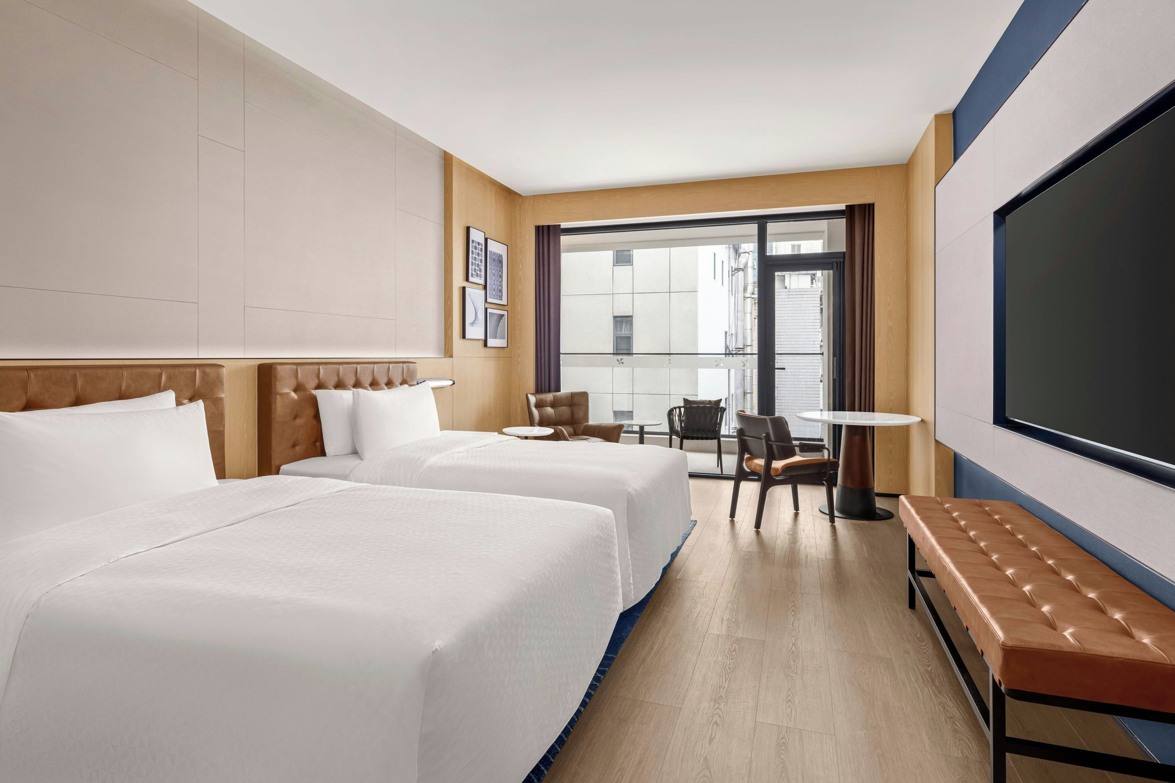 Habitación de hotel con dos camas, zona de estar con mesa y sillas, una gran pantalla de televisión y vistas al exterior.