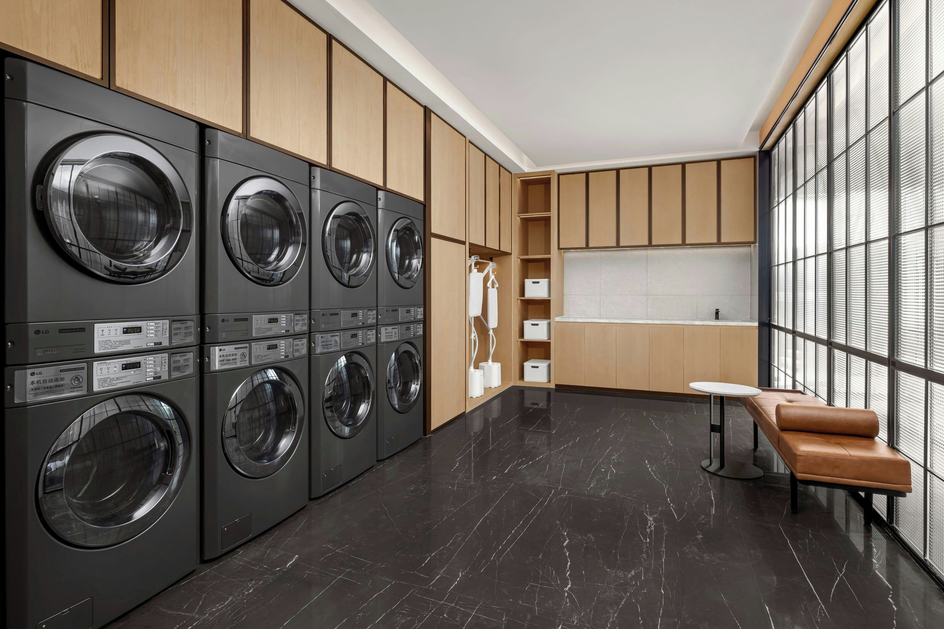 A modern laundry room with multiple LG commercial washers and dryers, wooden cabinets, a small seating area, and a large window providing natural light.