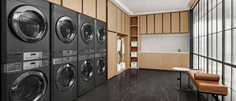 A modern laundry room with multiple LG commercial washers and dryers, wooden cabinets, a small seating area, and a large window providing natural light.