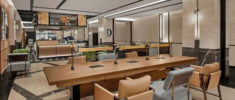 Un espace de restauration moderne dans un hôtel, avec une longue table en bois, des chaises en cuir et un bar à café en arrière-plan.