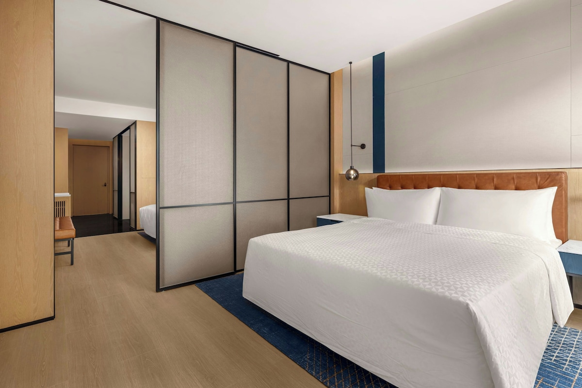 A hotel room with a large bed, a wooden headboard, a bedside table, and a view through a sliding door.