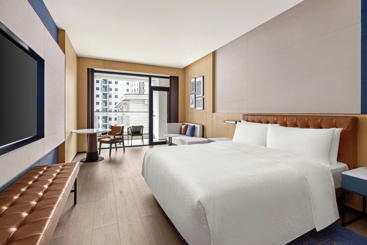 A hotel room with a large bed, a seating area with a table and chairs, a TV, and a view of the city through the window.