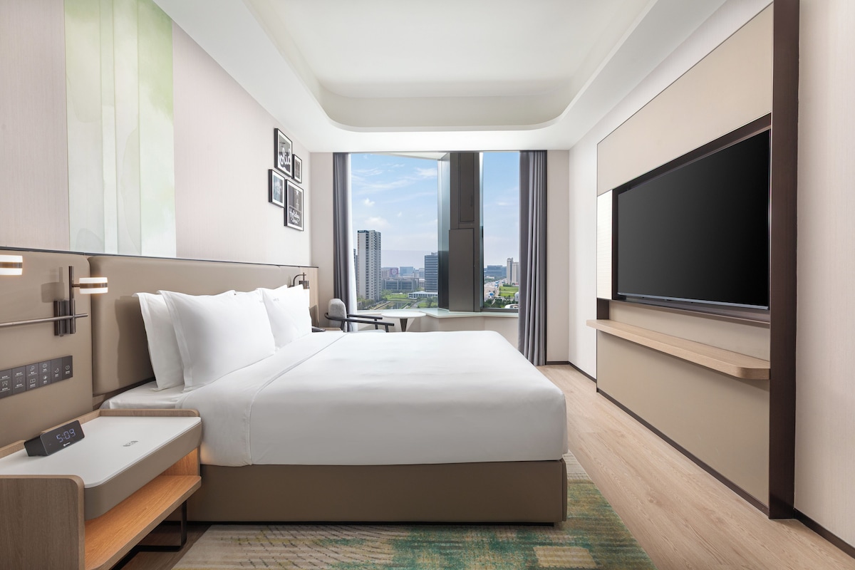 Una habitación de hotel moderna con una cama grande, un televisor de pantalla plana, un escritorio y vista a la ciudad.