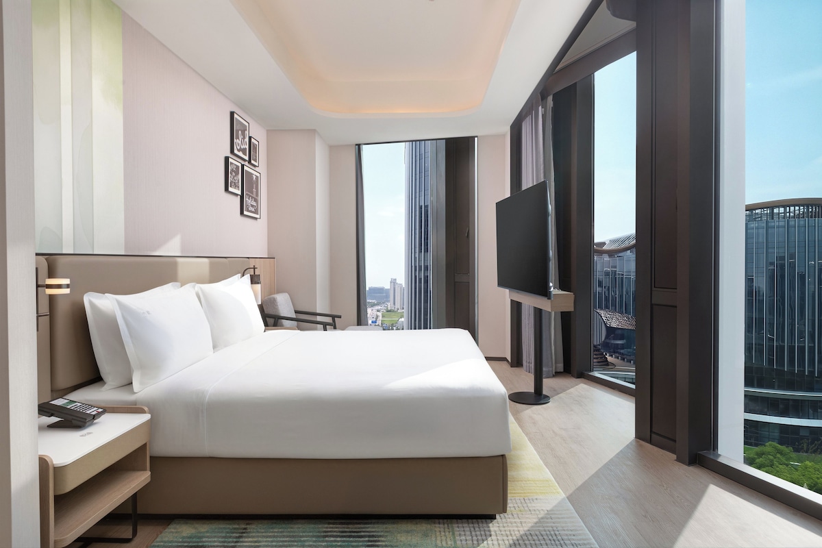 A modern hotel room with a large bed, a desk, a TV, and a city view.