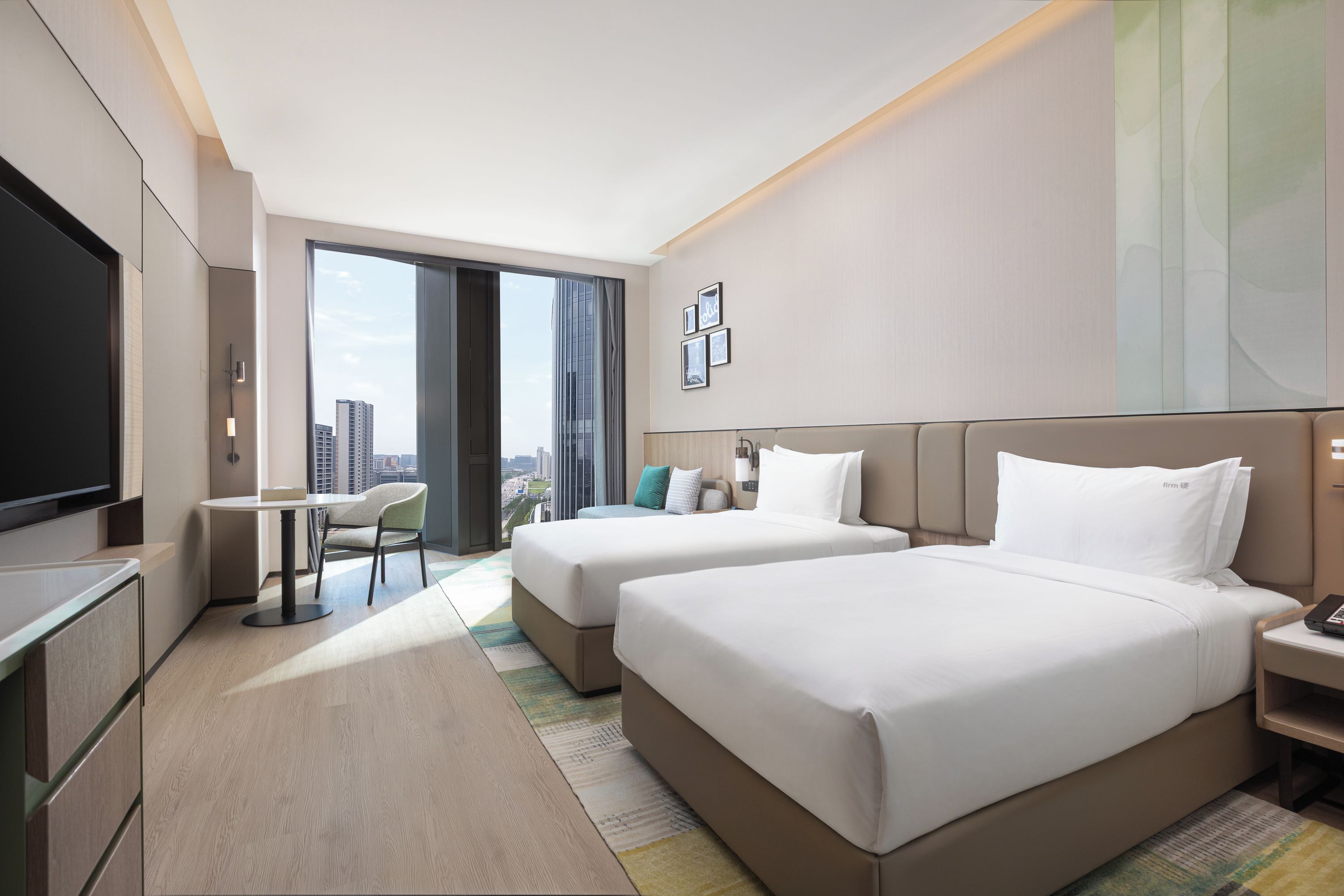A hotel room with two beds, a desk, a TV, and a view of the city.