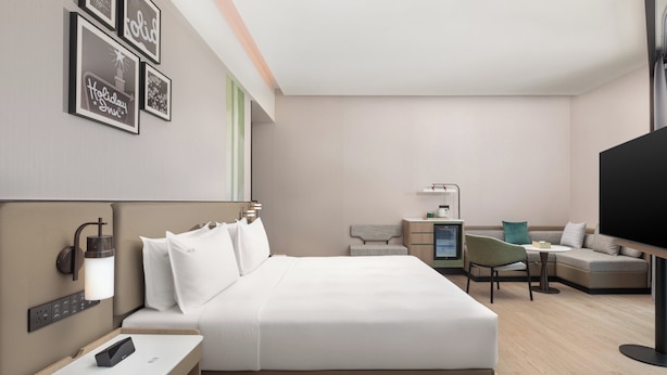 A modern hotel room with a bed, bedside table, lamp, TV, and a desk with a chair.