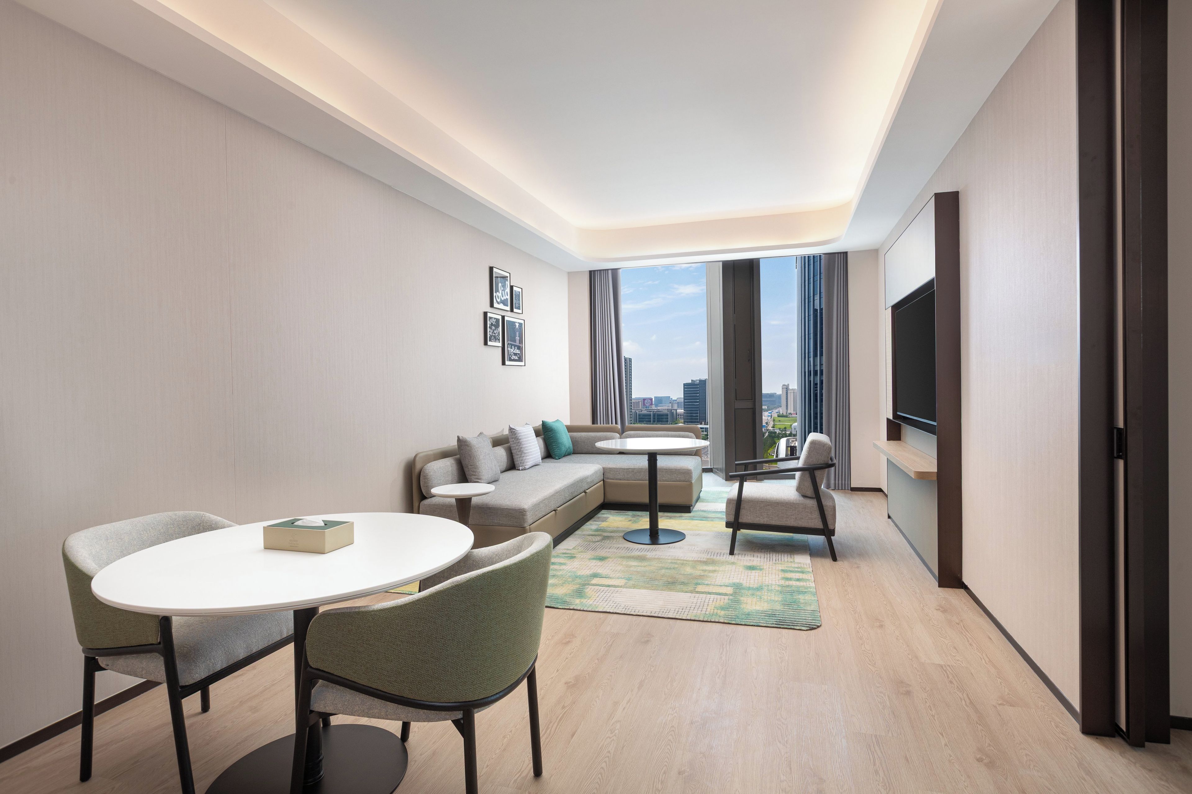 A modern hotel room with a sofa, a dining table, chairs, a TV, and a city view.