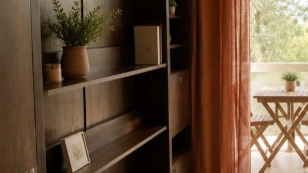 Una habitación con una estantería de madera, una mesita con una planta y vistas a través de una ventana con cortinas.